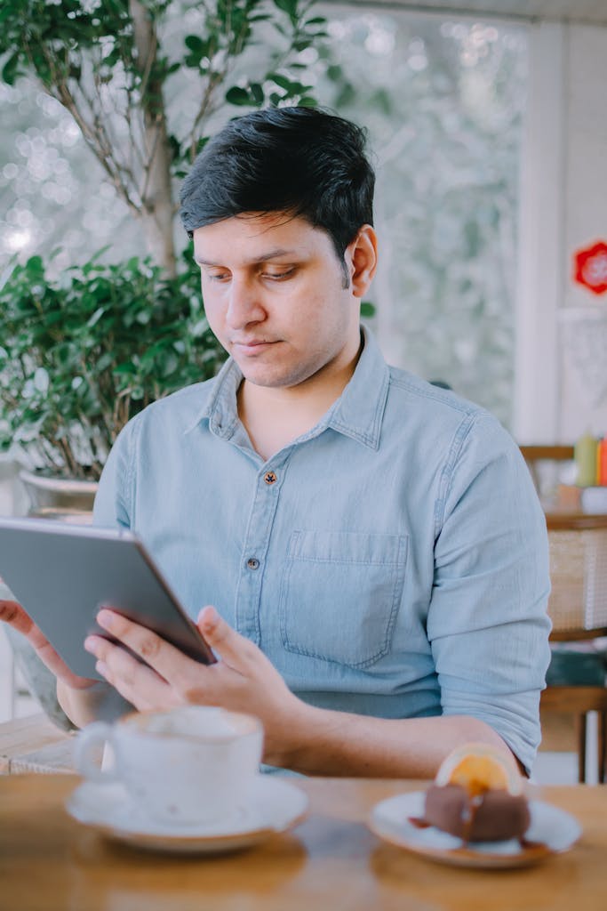 Adult man using a tablet in a relaxed cafe setting with coffee and dessert nearby.