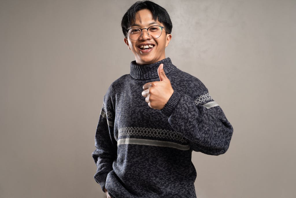 Asian man with eyeglasses smiling and giving a thumbs up, wearing a patterned sweater in a studio setting.