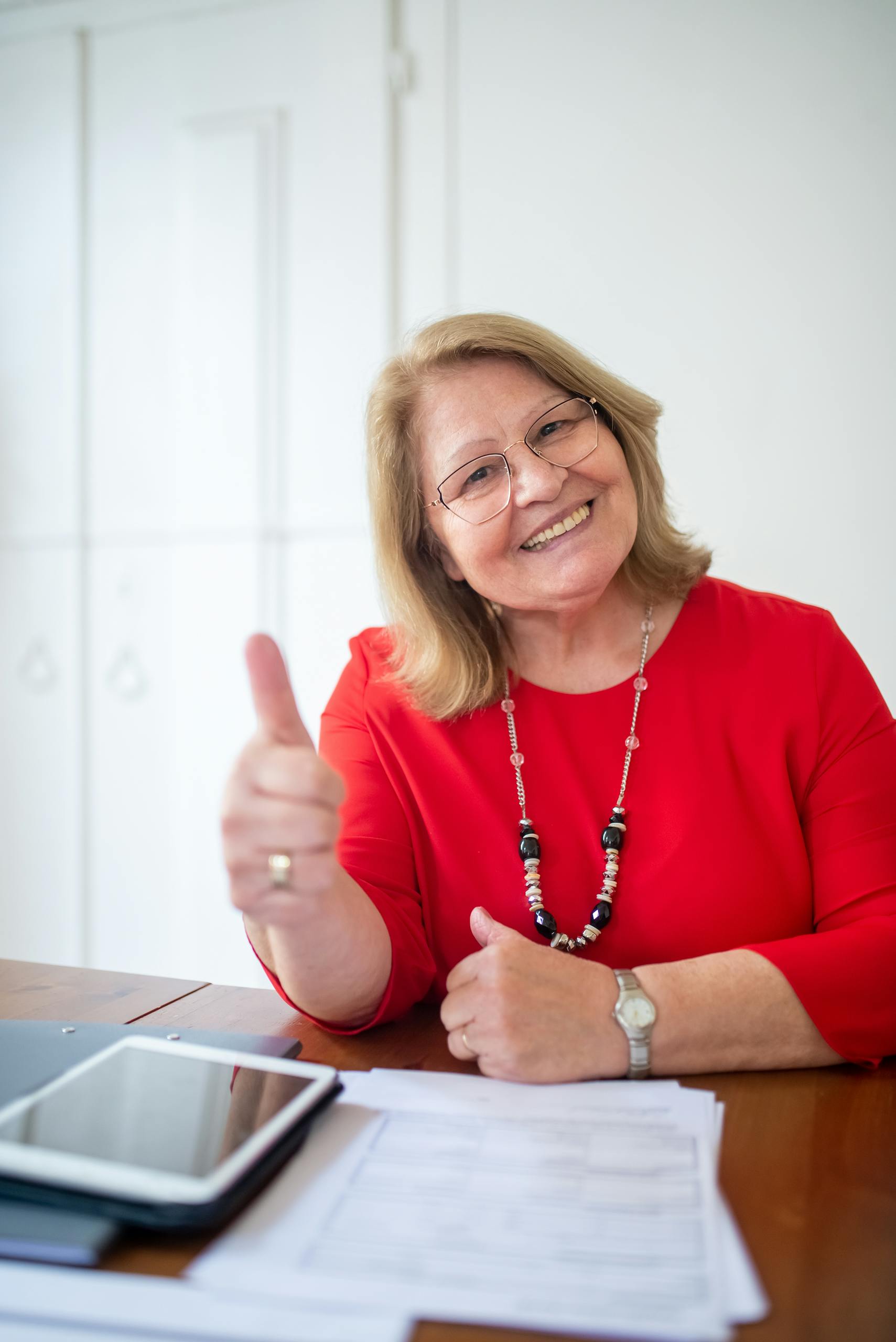Happy senior woman wearing glasses gives a thumbs up in a red shirt, displaying positivity.
