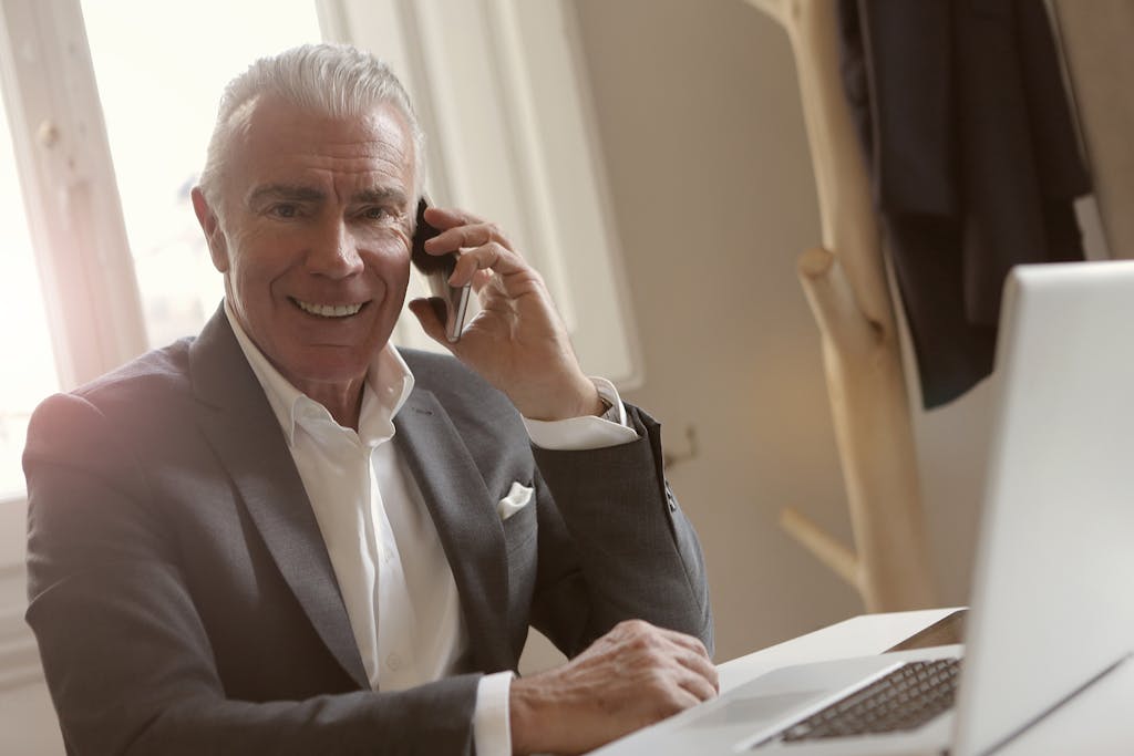 Senior man in a suit talking on a smartphone, working on a laptop in an office.