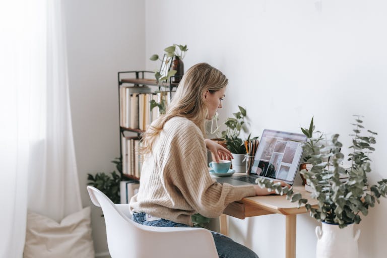 Side view of attentive female remote employee with cup of tea watching photo gallery on netbook screen at desk in house