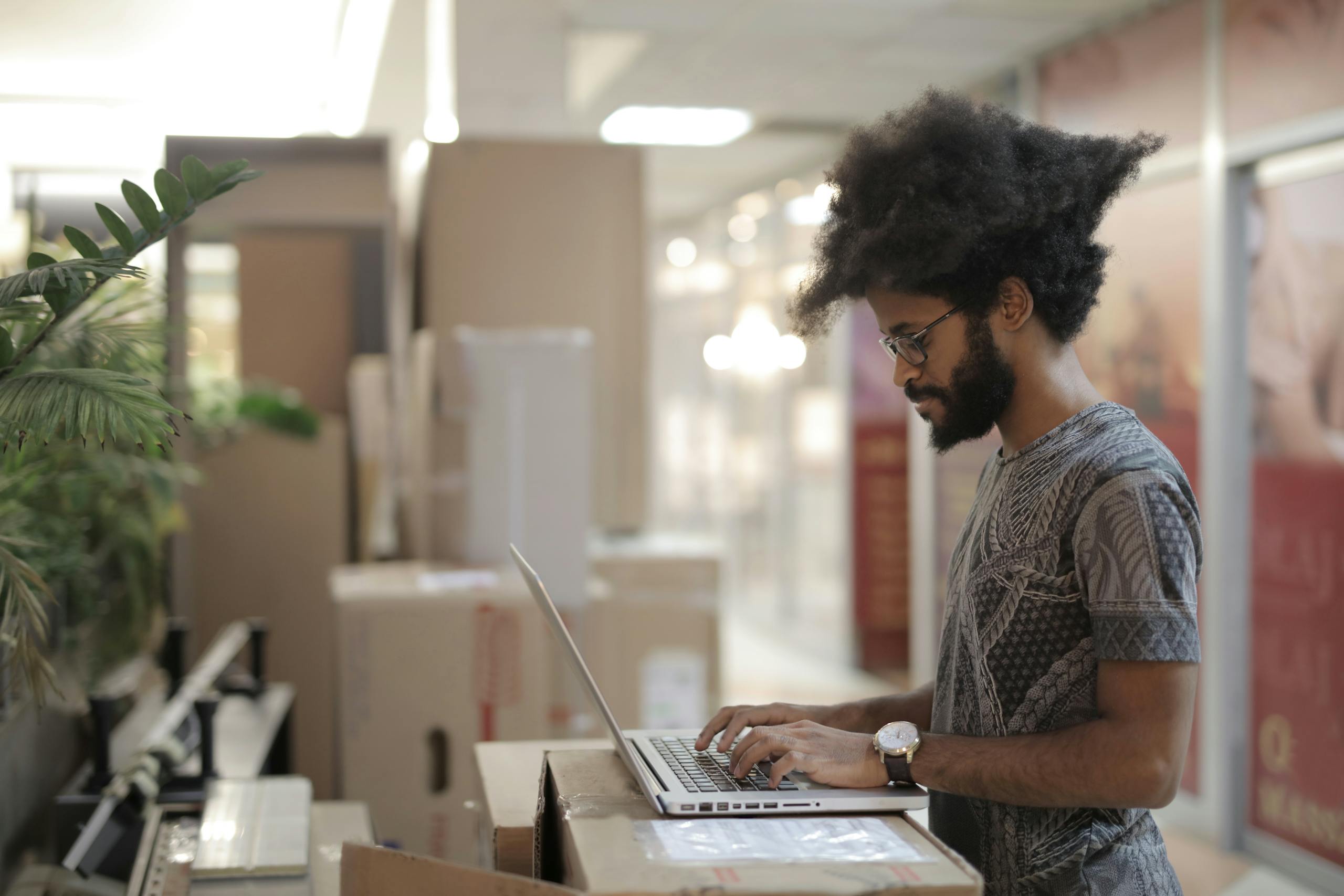 Side view of ethnic male with Afro hairstyle and in casual clothes with eyeglasses using laptop and thinking while working on project in modern workspace