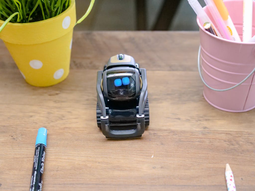 A small modern robot placed on a wooden desk surrounded by office supplies, showcases innovative technology.