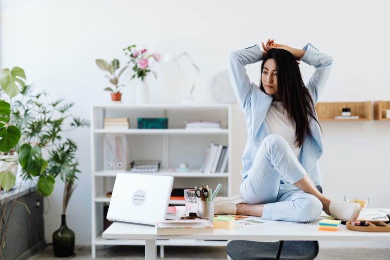 Asian businesswoman in a blue blazer sitting on a desk in a stylish office, creating a relaxed work atmosphere.